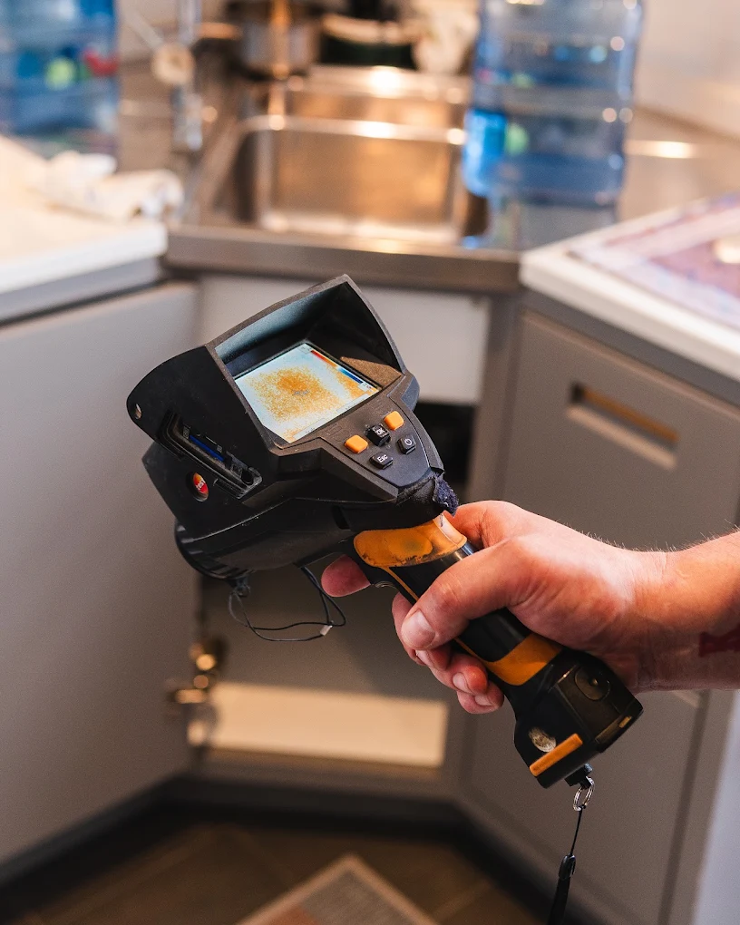 Thermal imaging camera scanning under a kitchen unit, its screen showing the heat pattern behind the surface