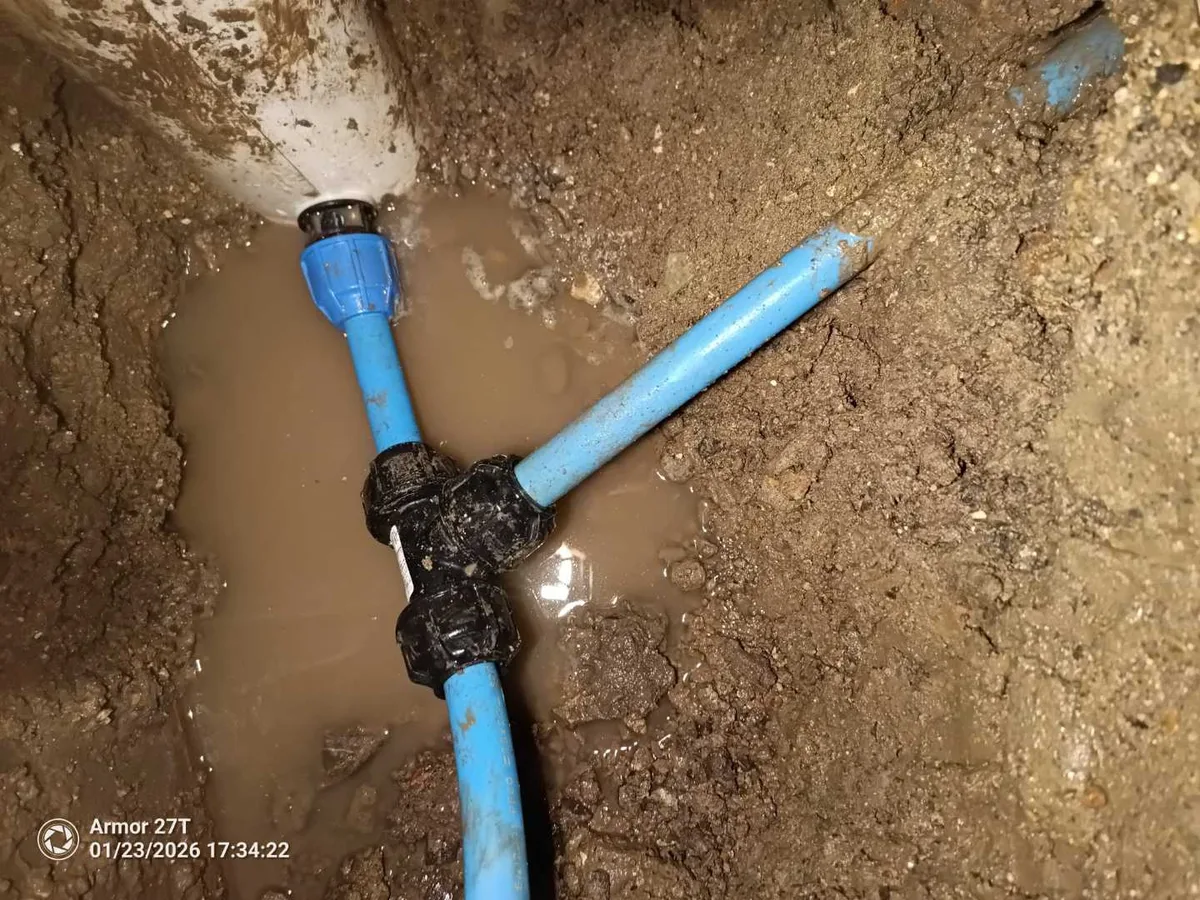 Blue MDPE pipe tee fitting exposed in a waterlogged excavation during an underground mains leak detection at a property in SN8