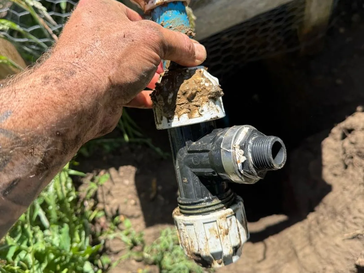 leak exposed at failed plastic tap assembly with corroded compression fittings held above excavation — leak located at