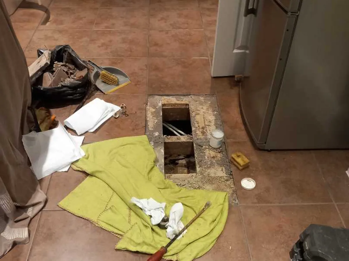 kitchen tiled floor with two tiles removed and floorboards lifted as repair in progress beneath fridge freezer